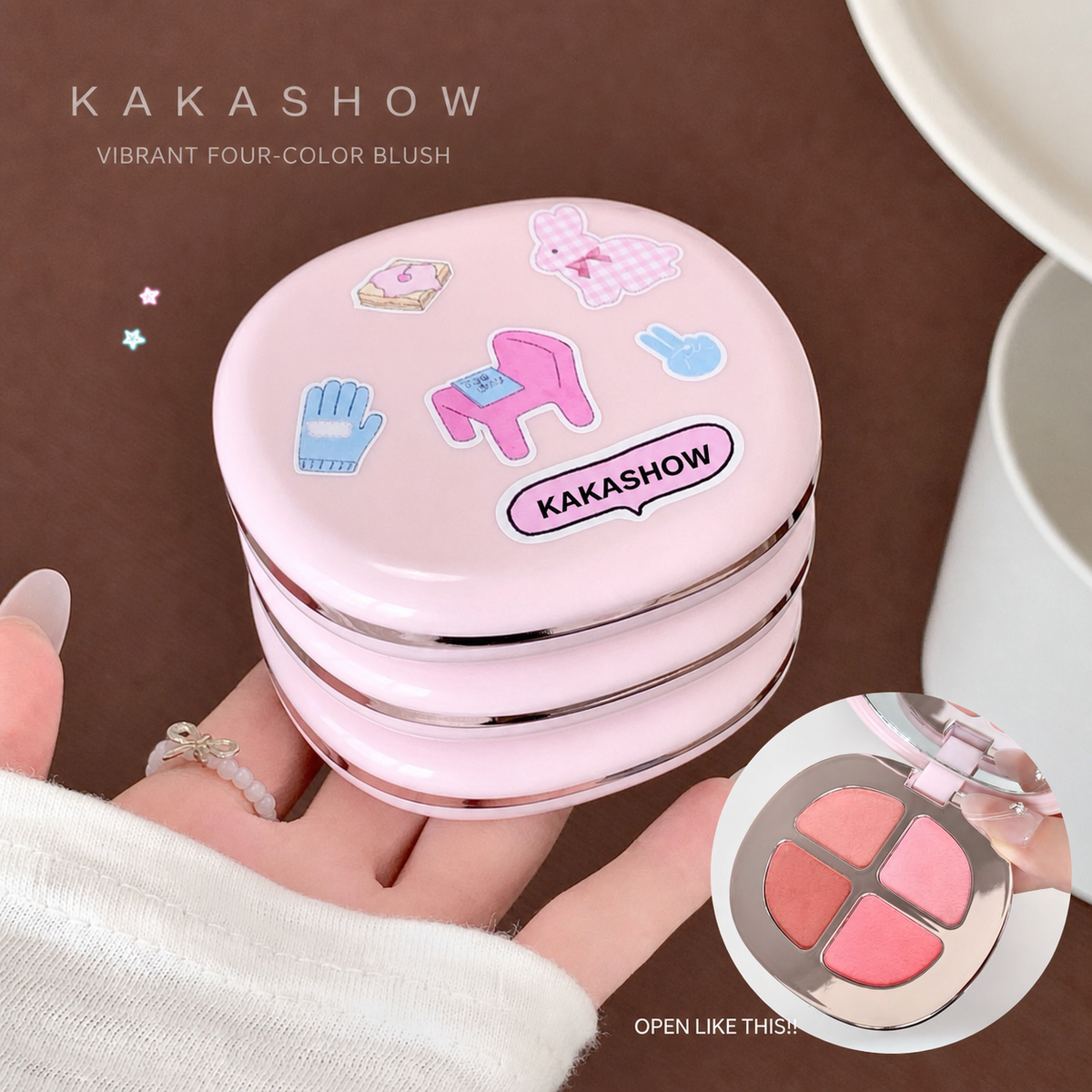 A pink round blush compact with decorative cartoon stickers on the lid, held in a person's hand. The compact has silver trim and a visible circular inset showing the opened four-color blush pans inside.