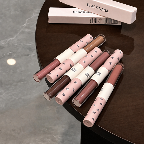Six lip gloss tubes from the brand BLACK NANA are arranged on a dark brown wooden table, with their white product boxes visible in the background.